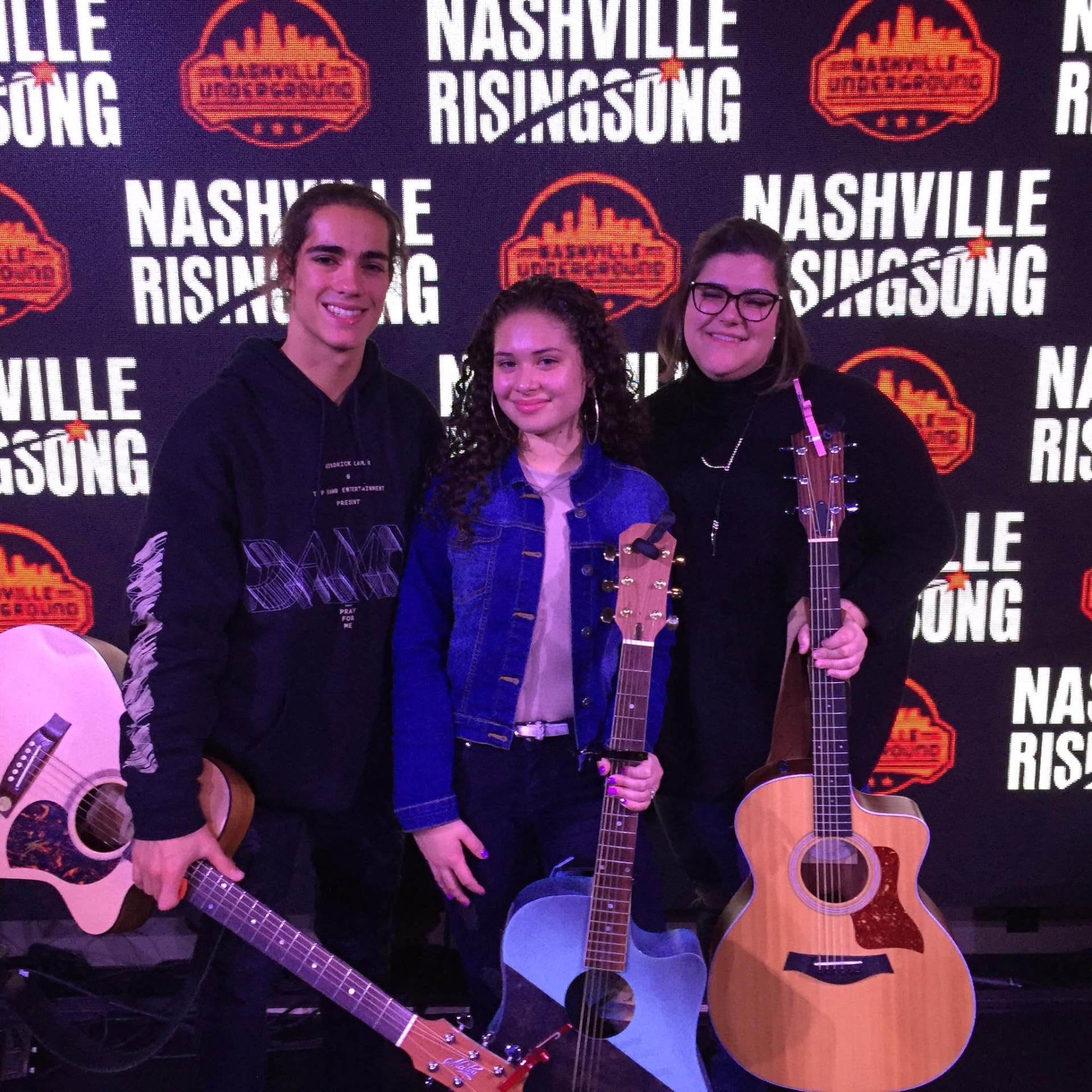 Three young songwriters with their guitars in front of the Nashville Rising Song backdrop